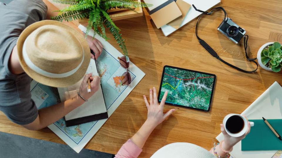  Two people plan a trip, surrounded by maps, a tablet displaying routes, a camera, and a coffee cup on a wooden table.