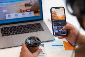 A person holds a coffee cup while browsing travel destinations on a smartphone, with a laptop displaying a beach background nearby.