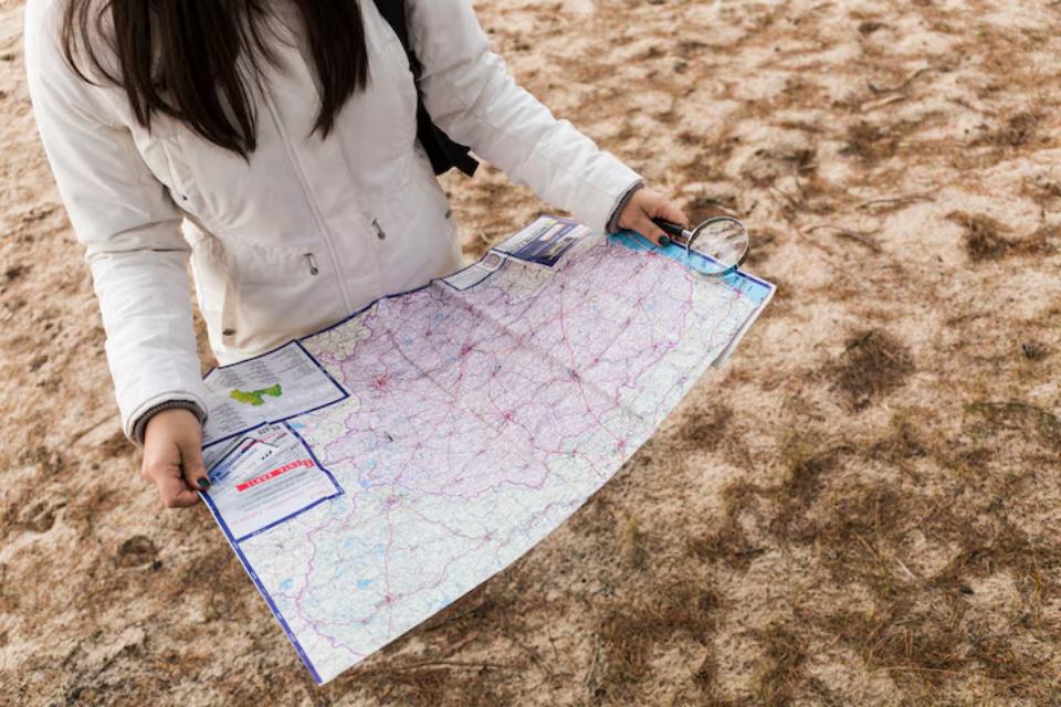 A person in a white jacket holds a large map and a magnifying glass, standing on sandy ground.