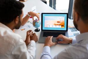 Two people are discussing vacation options while looking at a travel website on a laptop, with a globe and brochures on the table.