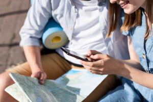 A young couple sits outdoors, one looking at a smartphone while the other holds a map, suggesting a travel planning moment.
