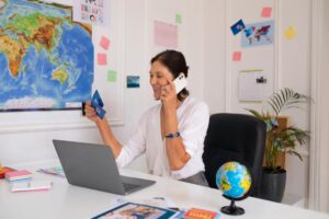 A woman in a white blouse talks on a phone while holding a credit card, seated at a desk with a world map and a globe.