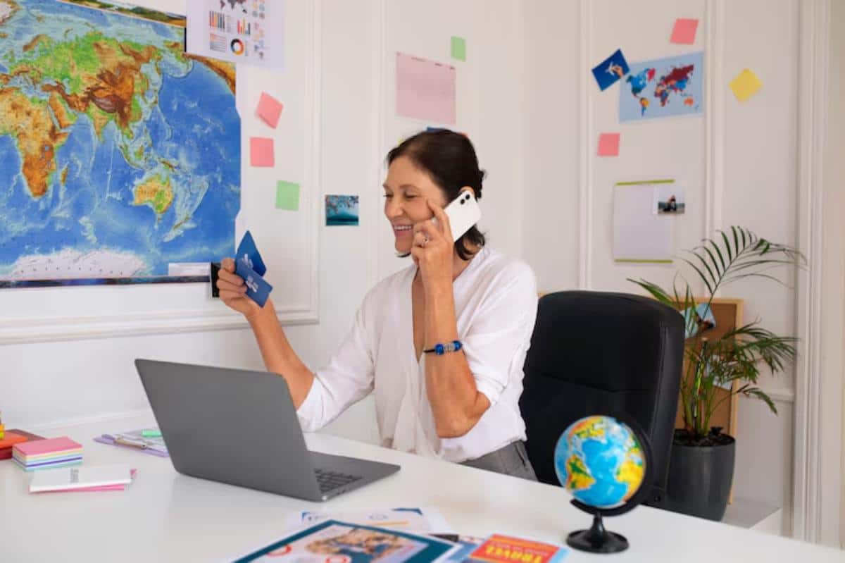 A woman in a white blouse talks on a phone while holding a credit card, seated at a desk with a world map and a globe.