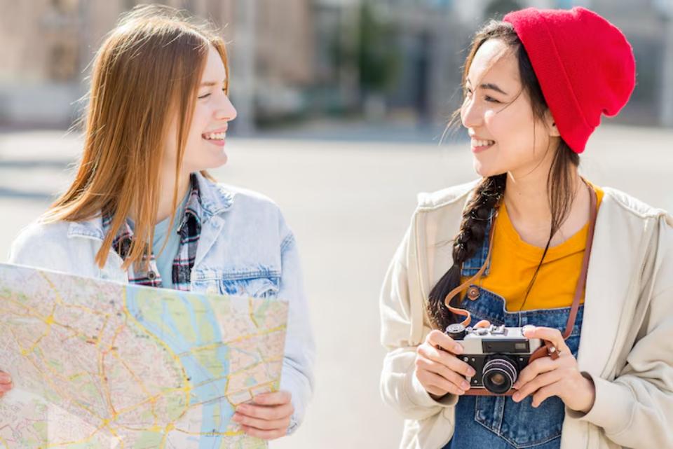 Two friends stand outdoors, one holding a map and the other a camera, sharing a moment while exploring a city.