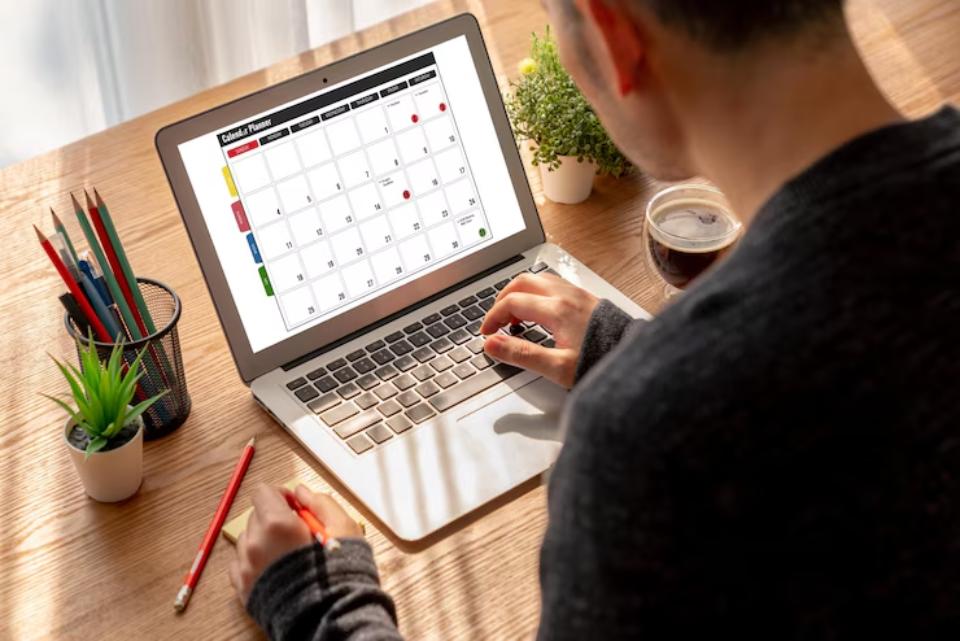  A person uses a laptop on a wooden desk, viewing a colorful calendar planner, surrounded by greenery and stationery. A coffee cup is nearby.