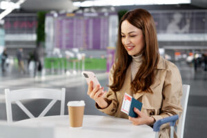 A woman in a beige trench coat sits at an airport table, holding her smartphone and a travel passport with a boarding pass beside a coffee cup.