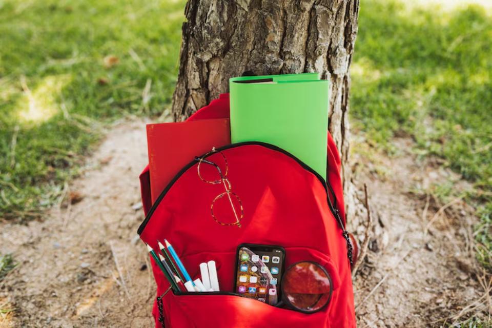  A red backpack resting against a tree, filled with notebooks, pens, glasses, and a phone, set on grass.