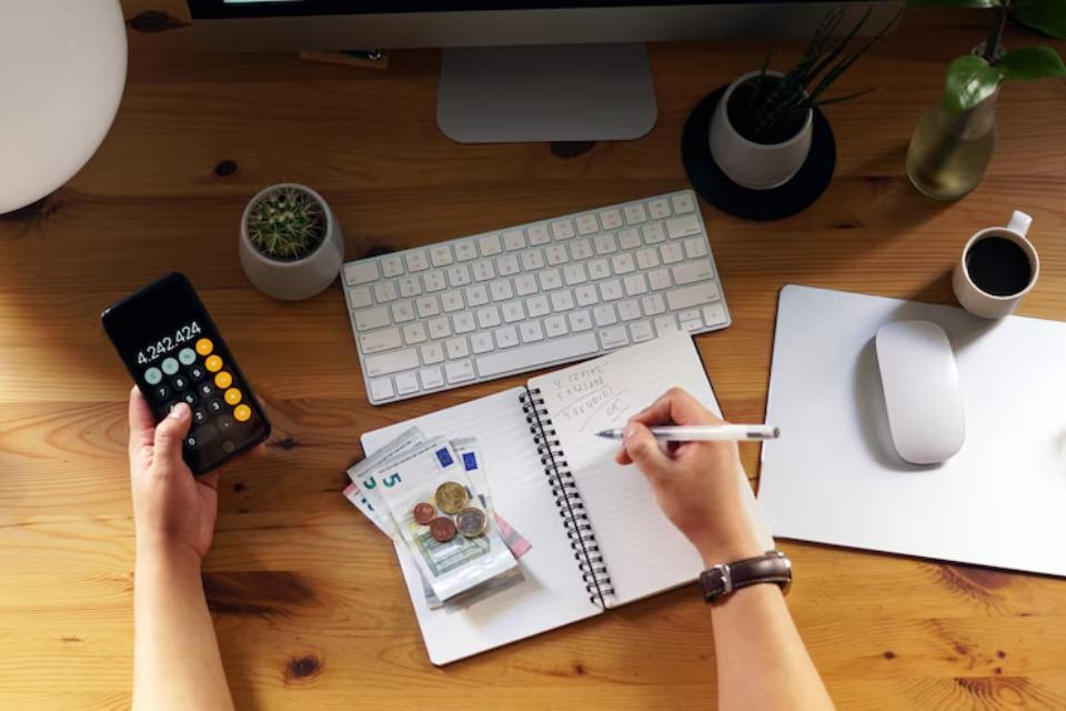  A desk scene showing a hand using a calculator on a smartphone, with a notebook, cash, coffee, and computer keyboard in view.