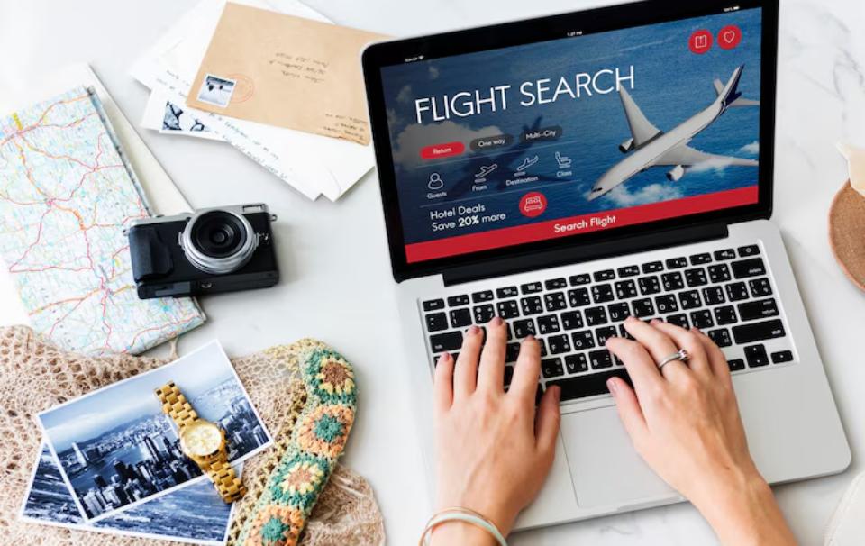A person searches for flights on a laptop surrounded by travel items, including a camera, postcards, a watch, and a map.