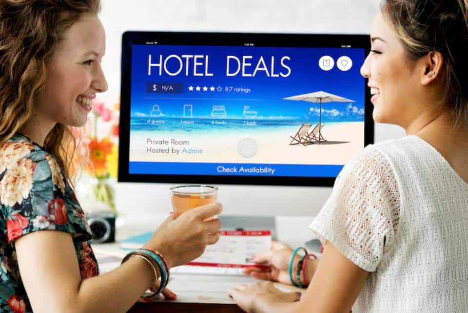 Two women are viewing hotel deals on a computer screen while one holds a drink and another has a booking document.