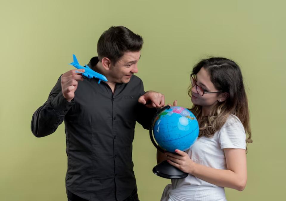  A couple engaging in travel planning, one holds a blue toy airplane, while the other points at a globe, set against a muted green background.