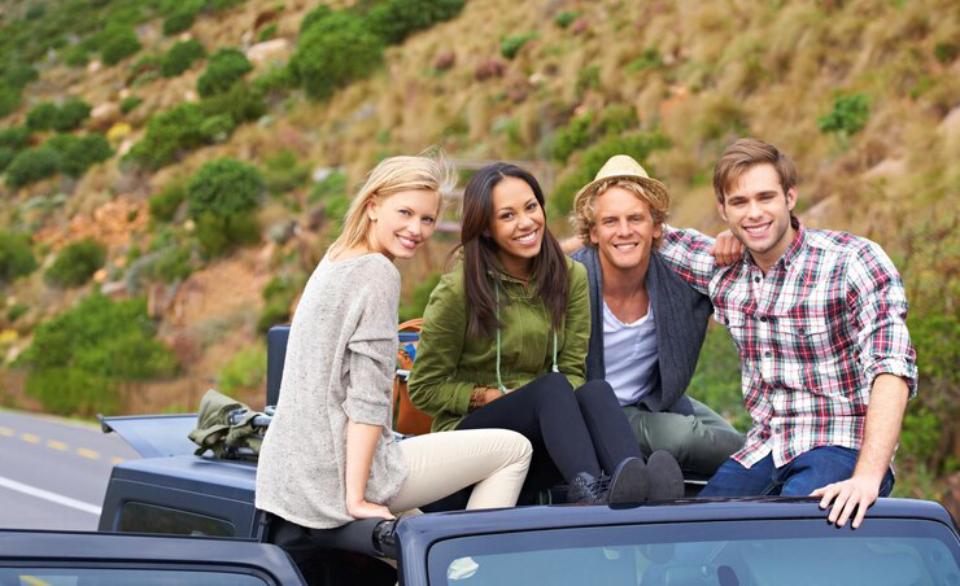  A group of four friends sitting on a car roof, enjoying a scenic roadside view with greenery and hills in the background.