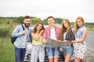 A group of five young adults stands by a car, studying a map in a green landscape on a cloudy day.