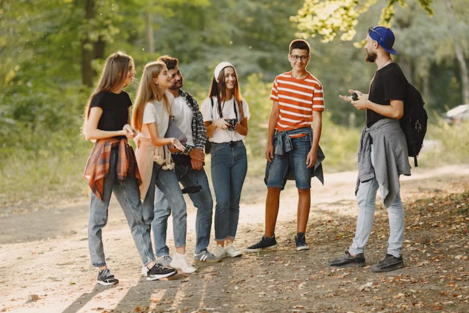  A group of six young people stands on a dirt path in a forest, chatting and enjoying a sunny day, surrounded by greenery and autumn leaves.