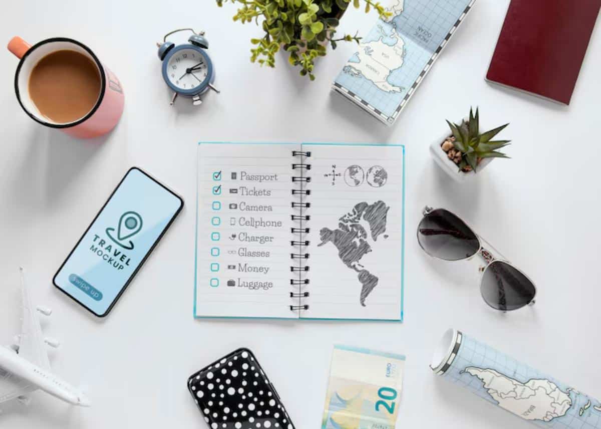 A flat lay of travel essentials: coffee, phone, notebook with a checklist, money, sunglasses, and a globe-themed travel mug.