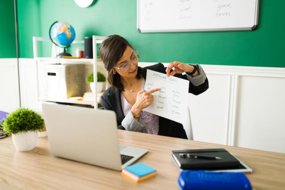 A woman points to a list of irregular verbs on a whiteboard in a bright classroom, with a laptop and stationery on her desk.