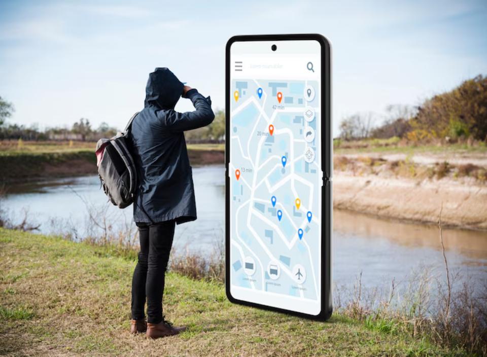  A person in a hoodie stands by a river, looking at a giant smartphone displaying a detailed map with various location markers.