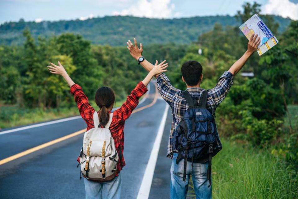 A young man and woman joyfully raise their arms on a sunny road, with a map in hand, surrounded by lush greenery and hills.