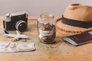 A travel-themed flat lay featuring a jar of coins labeled 'Travel', a vintage camera, cash, a toy airplane, passport, and a straw hat on a map.