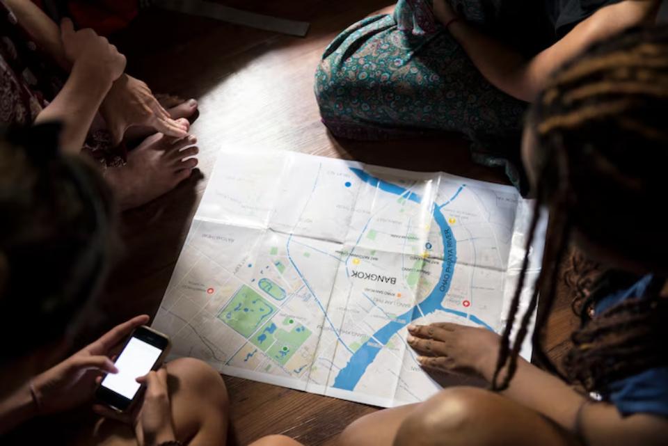 A group of people sitting around a folded map of Bangkok, one person holding a smartphone, with feet and hands visible.