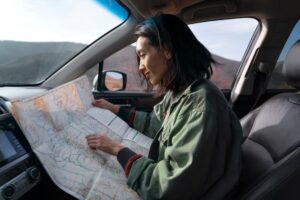 A person inside a car examines a detailed paper map, surrounded by a scenic view of mountains and blue skies.