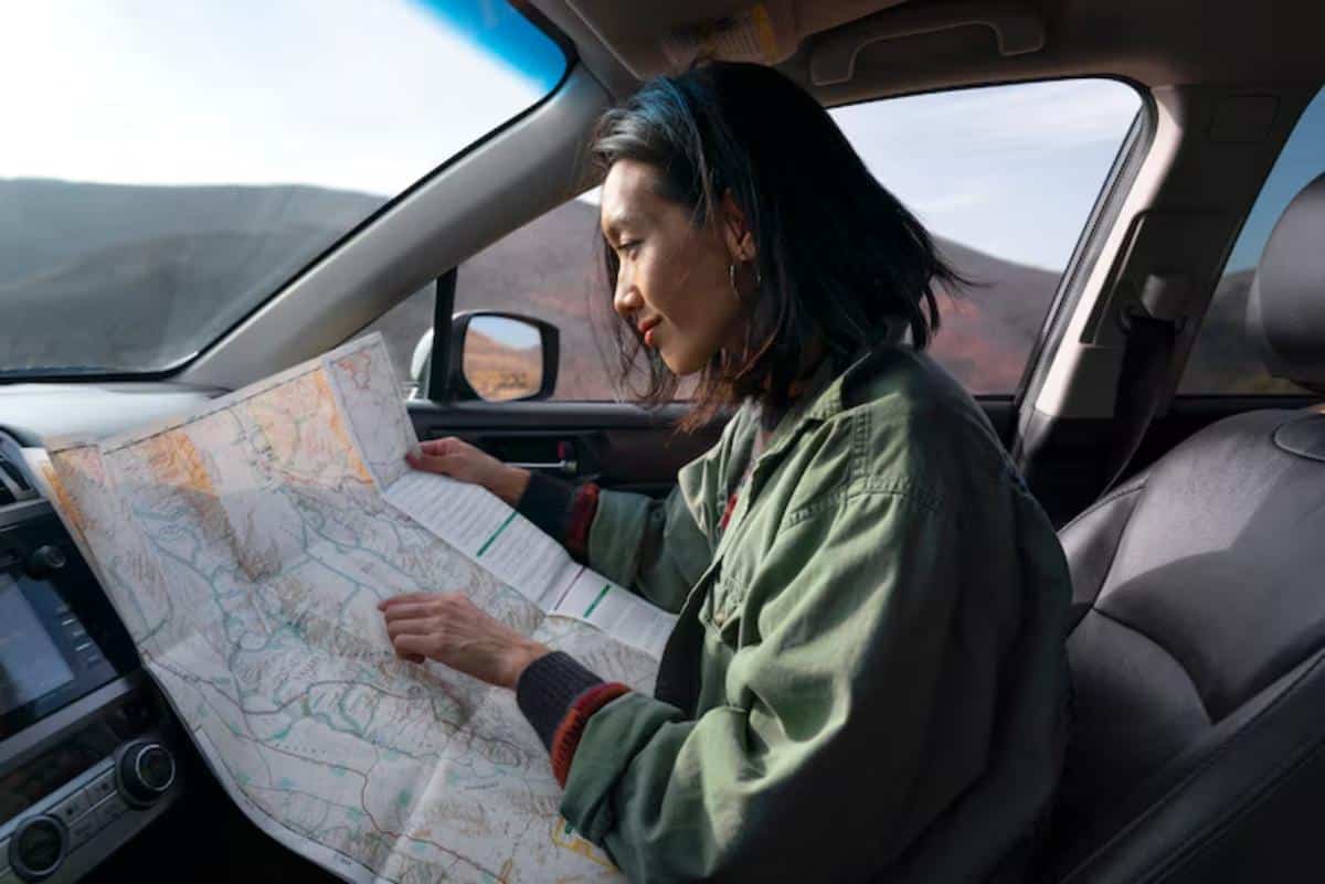 A person inside a car examines a detailed paper map, surrounded by a scenic view of mountains and blue skies.