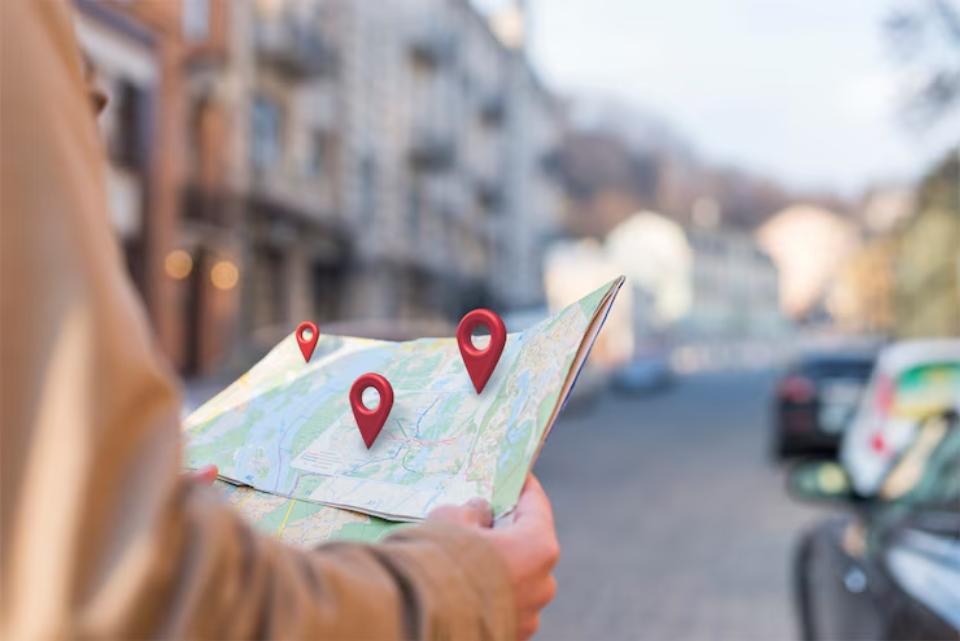 A person holds a map outdoors, with multiple red location pins indicating points of interest on the map. City buildings are blurred in the background.