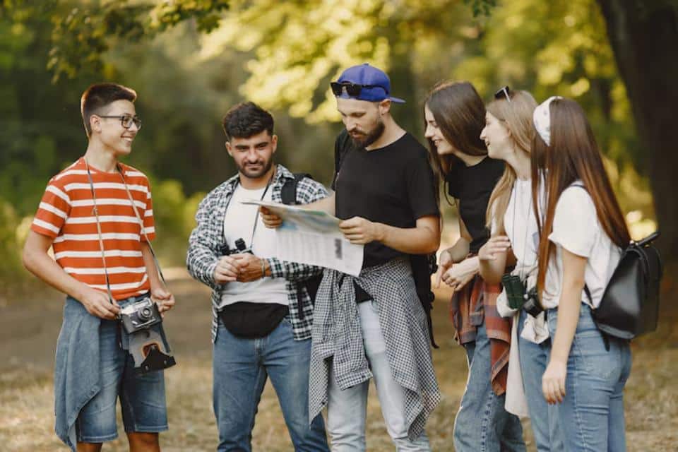 A group of young adults gathered in a forest, discussing a map while some hold cameras, enjoying a sunny day outdoors.