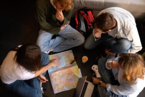 A diverse group of four young adults sits on the floor, examining a map and discussing travel plans, with a laptop and drinks nearby.