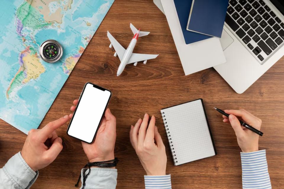  An overhead view of a travel setup: a smartphone, compass, airplane model, laptop, travel documents, and a notepad with a pen.