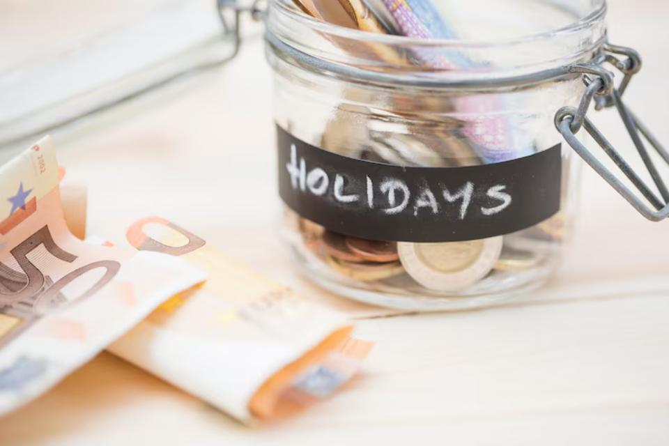  A glass jar labeled Holidays filled with coins and banknotes, next to folded euro bills on a light wooden surface.