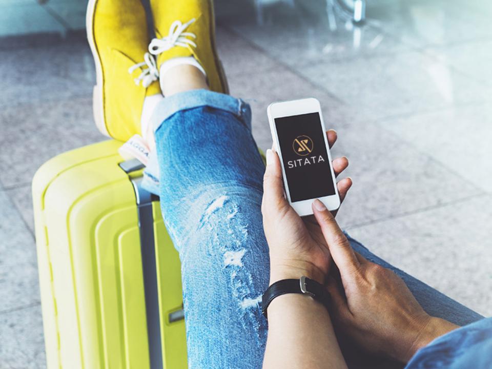  A person in yellow shoes relaxes with a smartphone displaying the SITATA app, beside a bright yellow suitcase in an airport lounge.