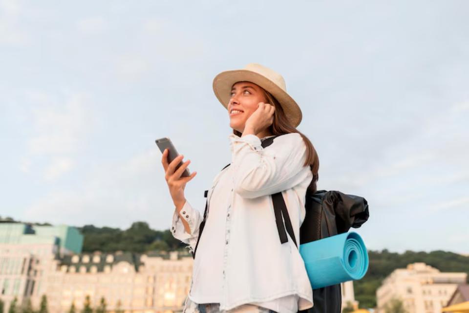  A young woman in a white shirt and straw hat holds a smartphone, with a black backpack and a blue yoga mat slung over her shoulder.