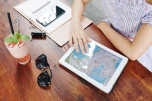 A hand points at a map on a tablet, surrounded by a smoothie, sunglasses, a phone, and a notebook on a wooden table.