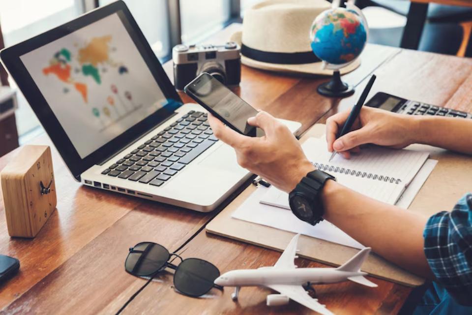  A person using a smartphone while planning a trip at a desk with a laptop, notebook, globe, camera, and airplane model.