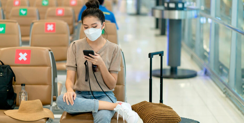 A person sits in an airport waiting area, holding a phone, with a suitcase and a hat beside them, surrounded by empty seats marked with signs.