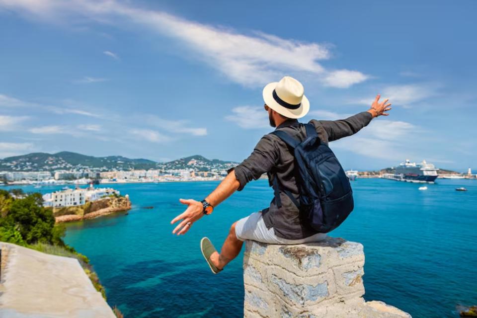  Man in a straw hat and backpack sitting on a stone ledge, arms outstretched, overlooking a vibrant blue sea and coastal town.