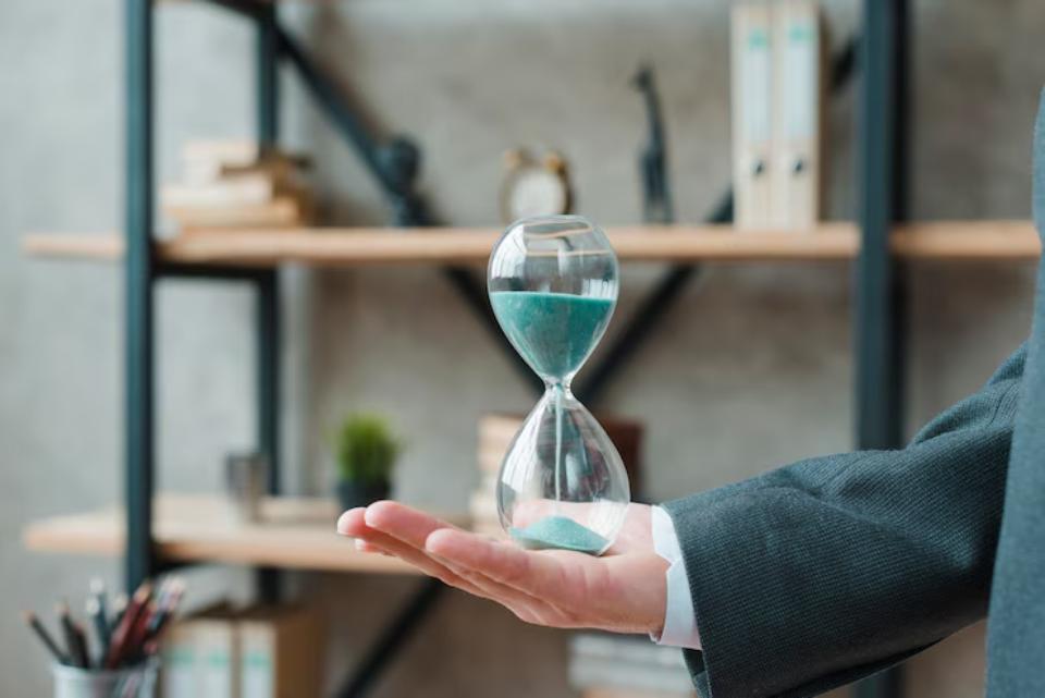  A person in a suit holds an hourglass with teal sand, symbolizing the passage of time, against a minimalist office background.