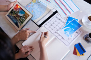 A top-down view of a workspace with a tablet displaying a photo, a notebook with travel plans, a calendar, and scattered documents.