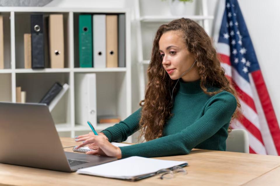  A woman with curly hair in a green sweater works on a laptop at a desk with paperwork, surrounded by bookshelves and an American flag.