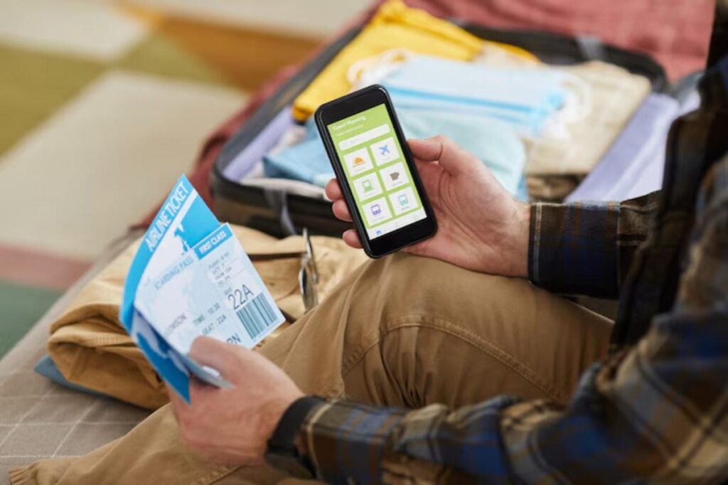 A traveler sits on a bed, holding an airline ticket and a smartphone displaying travel-related apps, surrounded by packed clothes.