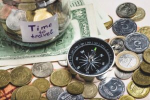 A jar labeled Time to travel is filled with coins, placed atop banknotes and surrounded by various coins and a compass.
