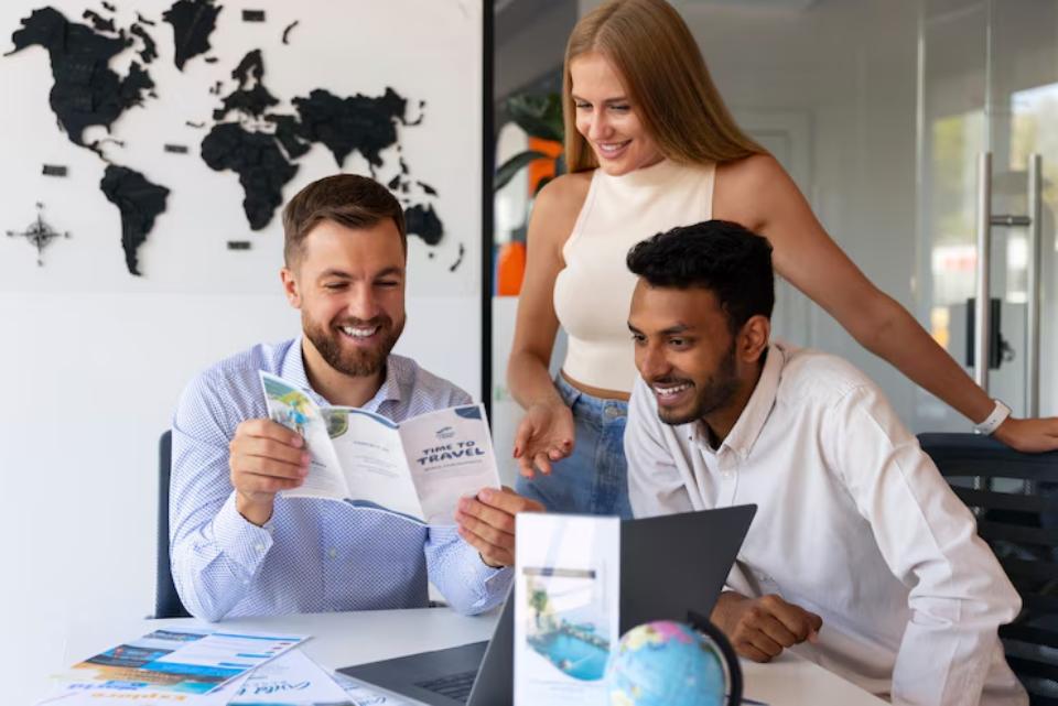 A group discusses travel plans at an office, with a world map on the wall and a laptop open on the table. Travel brochures are visible.