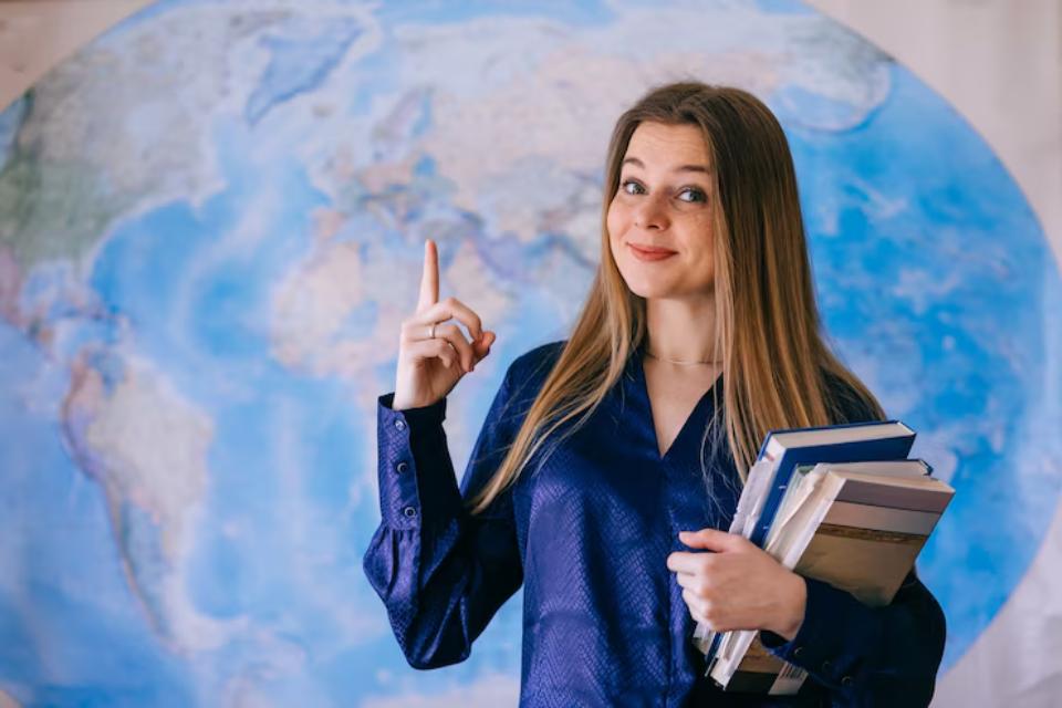  A woman in a blue blouse stands by a large globe, holding a stack of books and gesturing with one hand. 