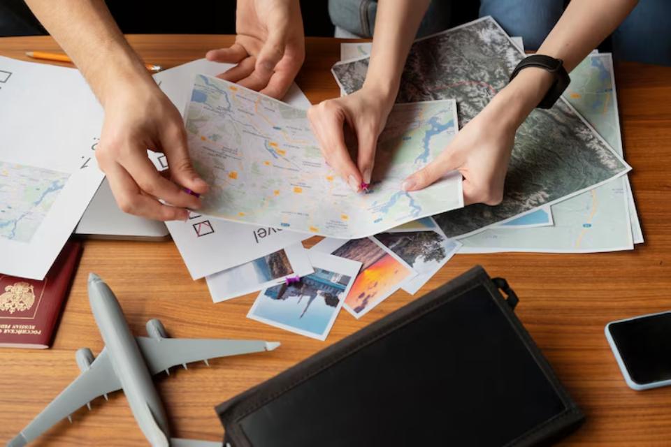  Two people are organizing various travel maps and photographs on a wooden table, with a passport and a model airplane nearby.