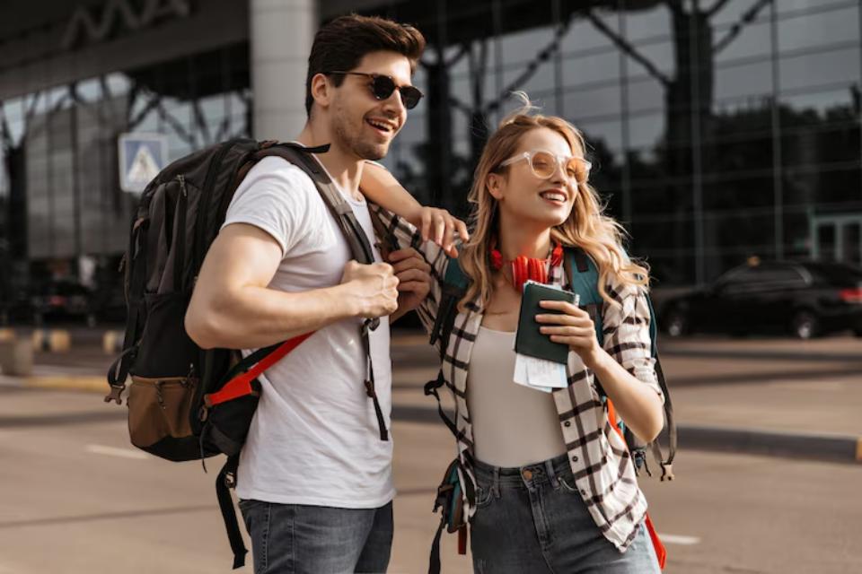  A young couple with backpacks stands outside an airport, engaged in conversation while holding a smartphone and travel documents.