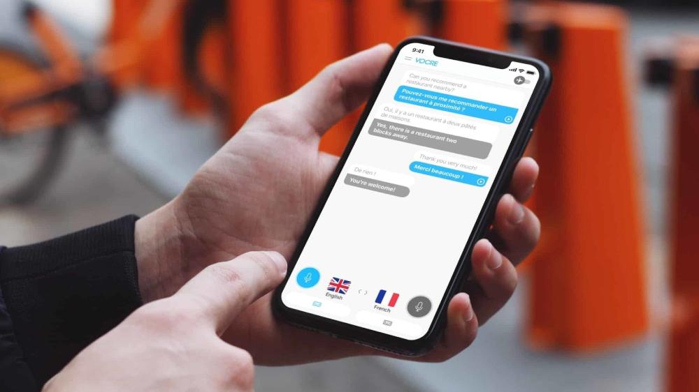  A person holds a smartphone displaying a language translation chat app with English and French messages about restaurant recommendations.