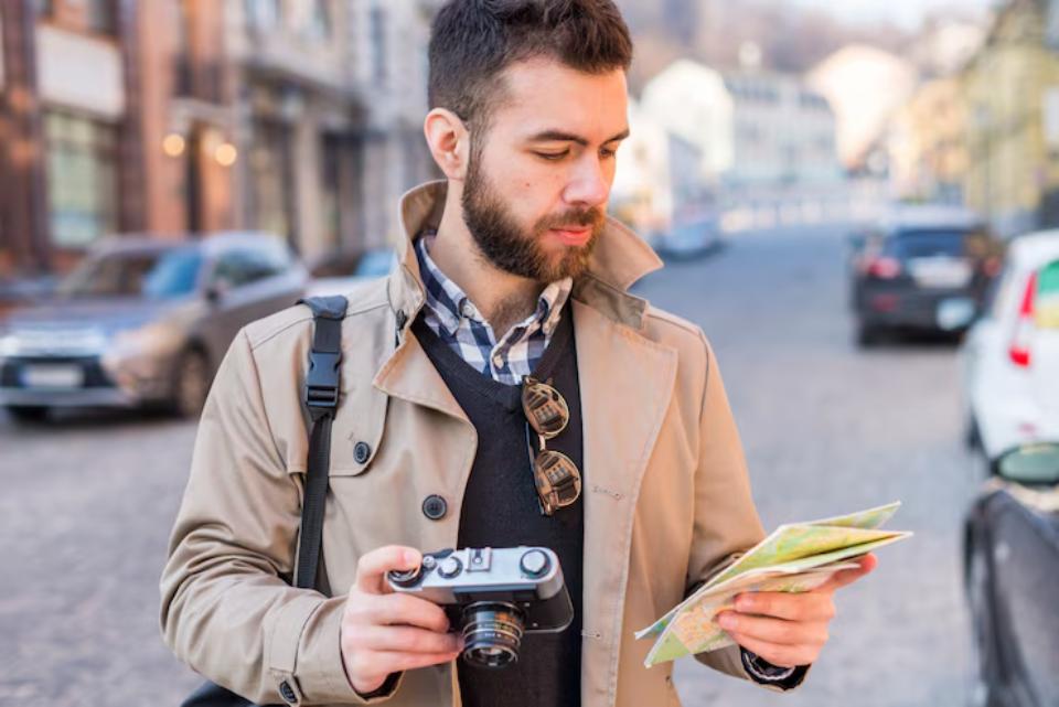A person in a beige trench coat holds a camera and a map while standing on a cobblestone street with parked cars in the background.